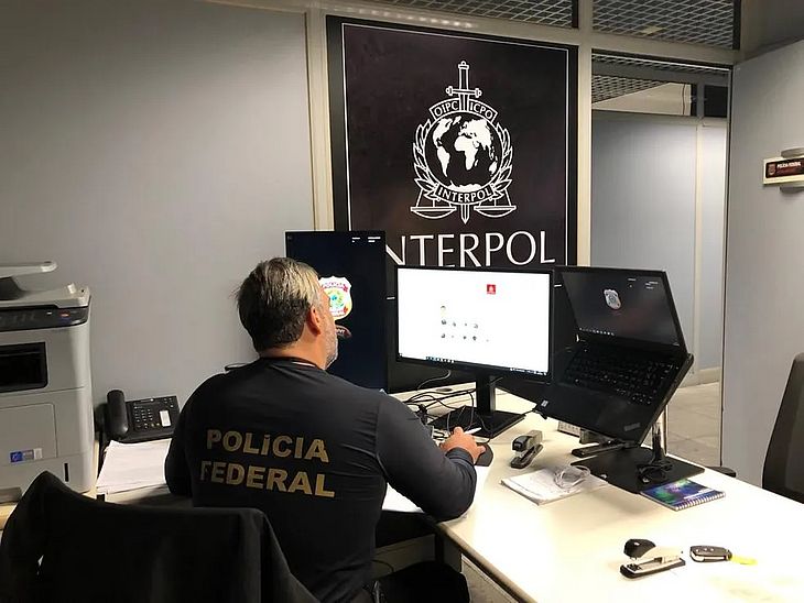 Imagem ilustrativa de um agente da Polícia Federal no escritório com a logo da Interpol